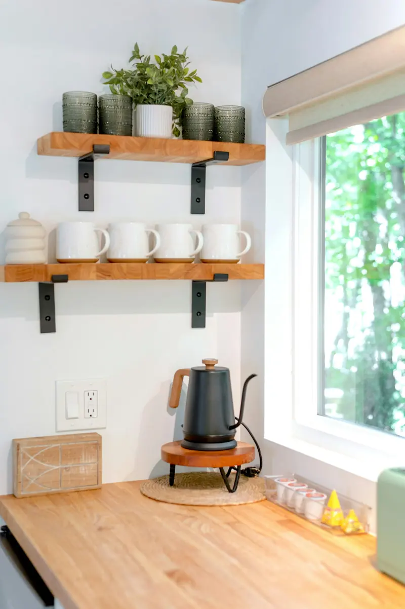 Sunlit kitchen corner: wooden open shelves with white mugs, black gooseneck electric kettle on a wooden stand, butcher-block counter, window with greenery.