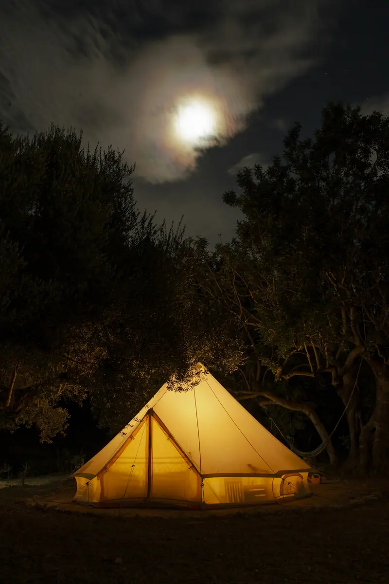 Glowing canvas bell tent at night among trees under a bright moon and cloudy sky.