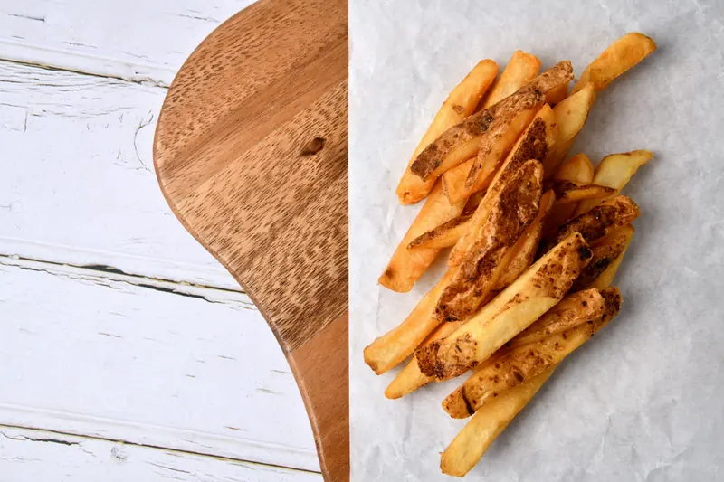 Golden seasoned fries on parchment beside a wooden serving board on a white-washed wood table, bright daylight.