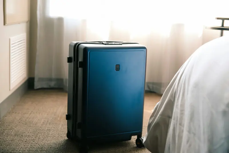 Blue hard-shell carry-on suitcase on carpet beside a hotel bed, backlit by sheer curtains.