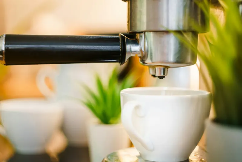 Espresso machine group head and black portafilter above a white cup, with potted plants and warm blurred home background.