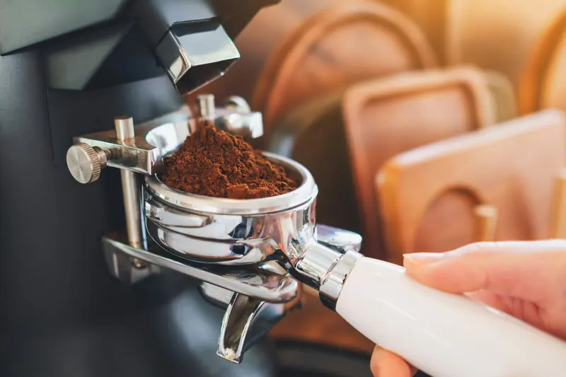 Freshly ground coffee dosing from a grinder chute into a portafilter held by a hand, warm blurred kitchen background.