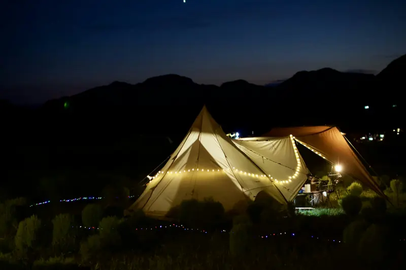 Canvas tents lit with warm string lights at a campsite at blue hour, dark mountains and sky behind.