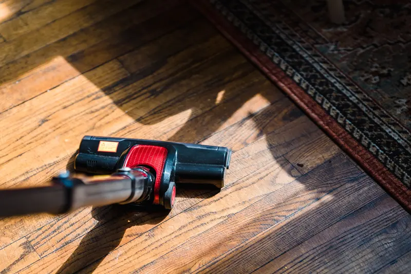 Close-up of a stick-vacuum floor head (black/red) on a sun-striped hardwood floor next to a patterned rug.